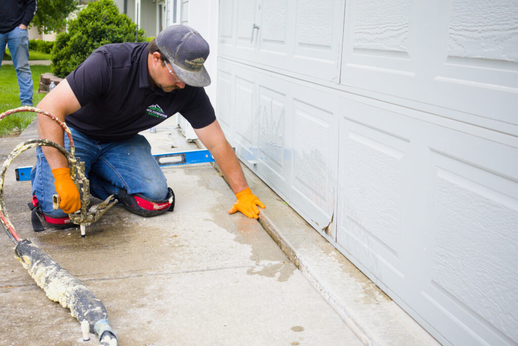Workman kneeling on a concrete driveway beside a white garage door, handling tools and a hose for a home repair project.